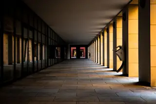 Illustration: A long, empty covered walkway lined with square pillars and glass windows in dramatic light.