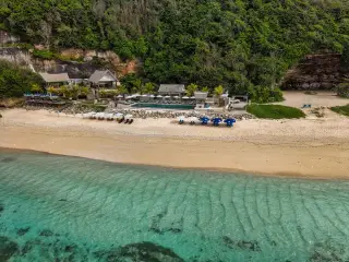 Illustration: Aerial view of a coastal beach club and resort near turquoise water and green cliffs.
