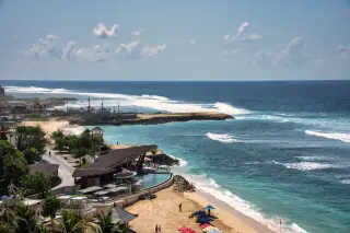Illustration: An elevated coastal view of a sandy beach, resort buildings, and waves rolling ashore under a blue sky.