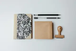 Illustration: A flatlay of a notebook, pens, brown paper block, and a wooden stamp on a white desk.