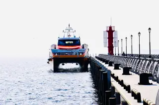 Illustration: A modern passenger ferry moored alongside a concrete pier with a red lighthouse.