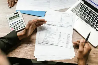 Illustration: Overhead view of hands discussing financial documents with a calculator and laptop on a wooden desk.