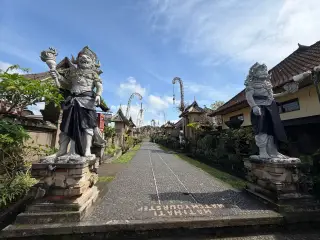 Illustration: Two stone guardian statues flank a paved empty village path lined with traditional houses and penjor poles.