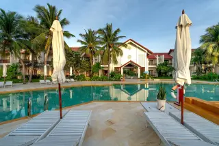 Illustration: Sun loungers and parasols by an empty swimming pool in front of a tropical resort building.