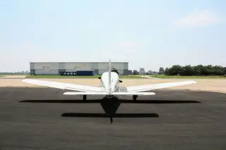 Illustration: Rear view of a small white airplane parked on an airport apron facing hangars.