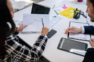 Illustration: Two people at an office desk review financial charts on paper, a tablet, and a laptop.