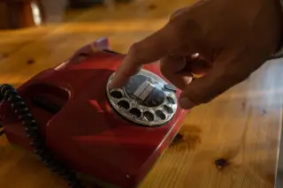 Illustration: A hand dials a vintage red rotary telephone resting on a wooden tabletop.