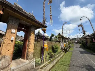 Illustration: Traditional stone gateways and tall penjor poles line a paved village street under a blue sky.