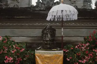 Illustration: A Balinese stone Ganesha statue sheltered by a ceremonial white umbrella flanked by pink flowers.