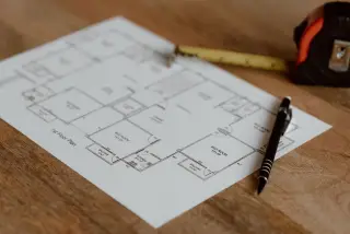 Illustration: A floor plan blueprint on paper beside a pen and tape measure on a wooden surface.