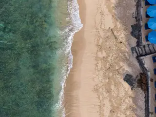 Illustration: An aerial view of waves lapping onto a sandy beach with blue umbrellas.