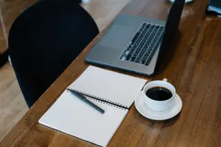 Illustration: A laptop, blank notebook, pen, and cup of coffee sit on a wooden desk.