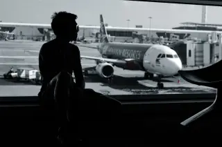 Illustration: Silhouette of a man sitting by an airport terminal window with an airplane on the tarmac outside.