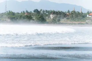 Illustration: Ocean waves rolling toward a hazy tropical coastline with buildings, palm trees, and distant hills.