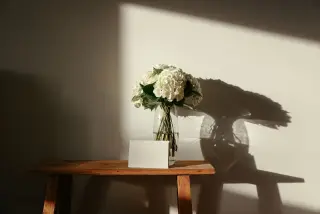 Illustration: White flowers in a glass vase and a blank card rest on a wooden bench against a wall.