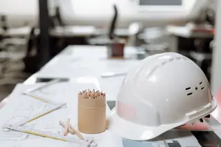 Illustration: A white hard hat, colored pencils, and architectural plans on an office desk.