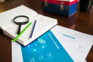 Illustration: A notebook with a magnifying glass, pencils, and business report sheets on a wooden desk.