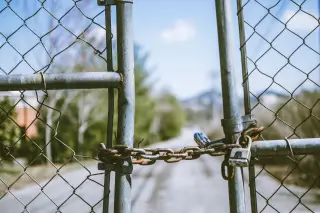 Illustration: A chain link gate fastened with a rusty chain and a padlock overlooking an empty road.