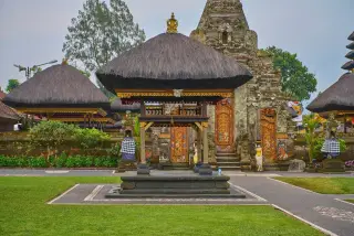 Illustration: A Balinese temple complex with thatched-roof pavilions surrounded by grass and stone paving.