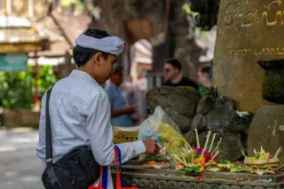 Illustration: A man in traditional Balinese attire places offerings near a stone monument with Balinese script.