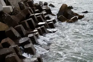 Illustration: Ocean waves crash against concrete breakwater tetrapods on a coastline.