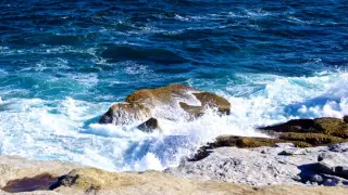Illustration: Ocean waves crashing against rocky shoreline boulders under bright sunlight.