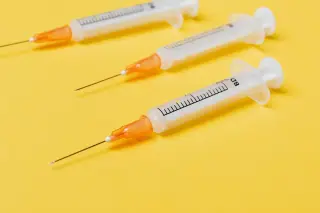 Illustration: Three medical syringes with bare needles lying on a bright yellow background.