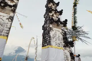 Illustration: Traditional Balinese temple gate decorated with fabric and penjor with a mountain in the background.