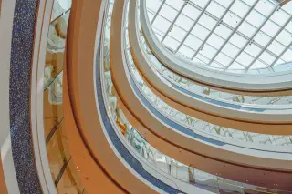 Illustration: Low-angle view of curved modern mall balconies beneath a glass skylight ceiling.