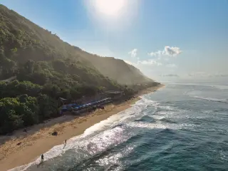 Illustration: Aerial view of a sunny tropical beach bordered by a forested cliff and ocean waves.