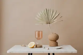 Illustration: A minimalist still life with an hourglass, lemon slice, and dried palm leaf in a vase on a marble table.