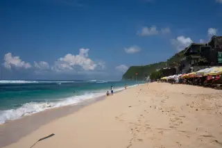 Illustration: People relaxing on a sunny sandy beach beside ocean waves and cliffside buildings.