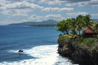 Illustration: A rocky coastal cliff with palm trees and a small gazebo overlooking the blue ocean.