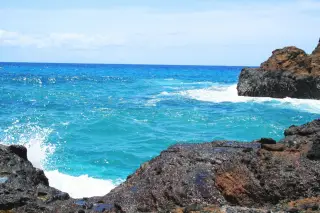 Illustration: Turquoise ocean waves crashing against dark rocky outcrops under a clear sky.