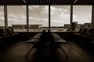 Illustration: Empty airport terminal seats facing large windows looking out onto an airfield and buildings.