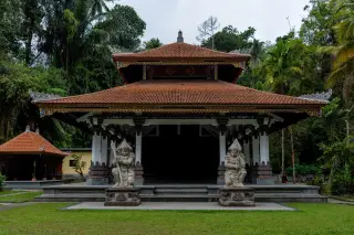 Illustration: A traditional Balinese pavilion flanked by guardian stone statues in a green garden.