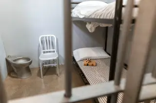 Illustration: View through metal bars into a clean prison cell with bunk beds, a chair, and a toilet.