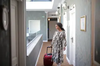 Illustration: A woman walking with a red rolling suitcase along a hotel hallway.