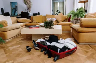 Illustration: An open red suitcase with clothes, shoes, and a hat lies on a parquet floor in a living room.