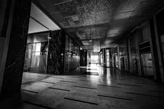 Illustration: Black and white shot of an empty indoor building hallway with light at the far end.