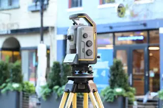 Illustration: A modern electronic total station mounted on a yellow tripod on a city street.