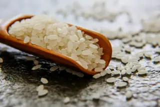 Illustration: Close-up of raw white rice grains spilling from a small wooden scoop onto a dark surface.