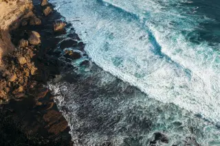 Illustration: Overhead view of ocean waves breaking against a rocky shoreline and cliff base.