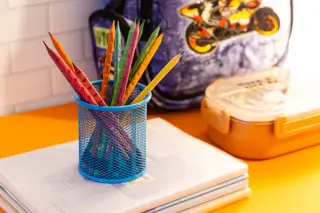 Illustration: Colored pencils in a blue mesh holder sit on notebooks beside a lunchbox and backpack.