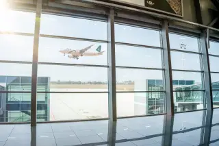 Illustration: An airplane taking off viewed through large glass windows of an airport terminal.
