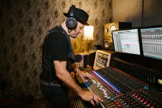 Illustration: A man wearing a hat and headphones operates a sound mixing console in a recording studio.