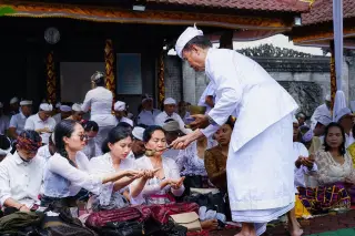 Illustration: A Balinese priest pours holy water onto the hands of seated worshippers in traditional attire.