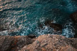 Illustration: A high-angle view of ocean waves and white foam washing against a rocky cliff.