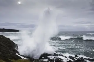 Illustration: A powerful sea wave sprays high against a rocky shore under an overcast sky.