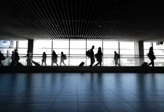 Illustration: Silhouettes of travelers walking through an airport terminal with large windows.